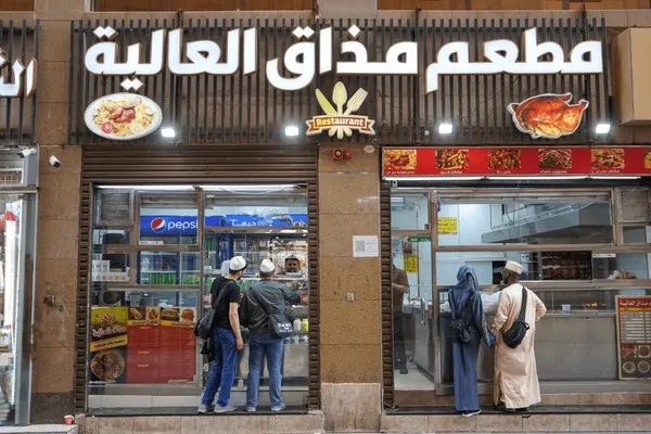 Customers ordering food at a restaurant in Saudi Arabia, with a sign displaying Arabic text and menu items.