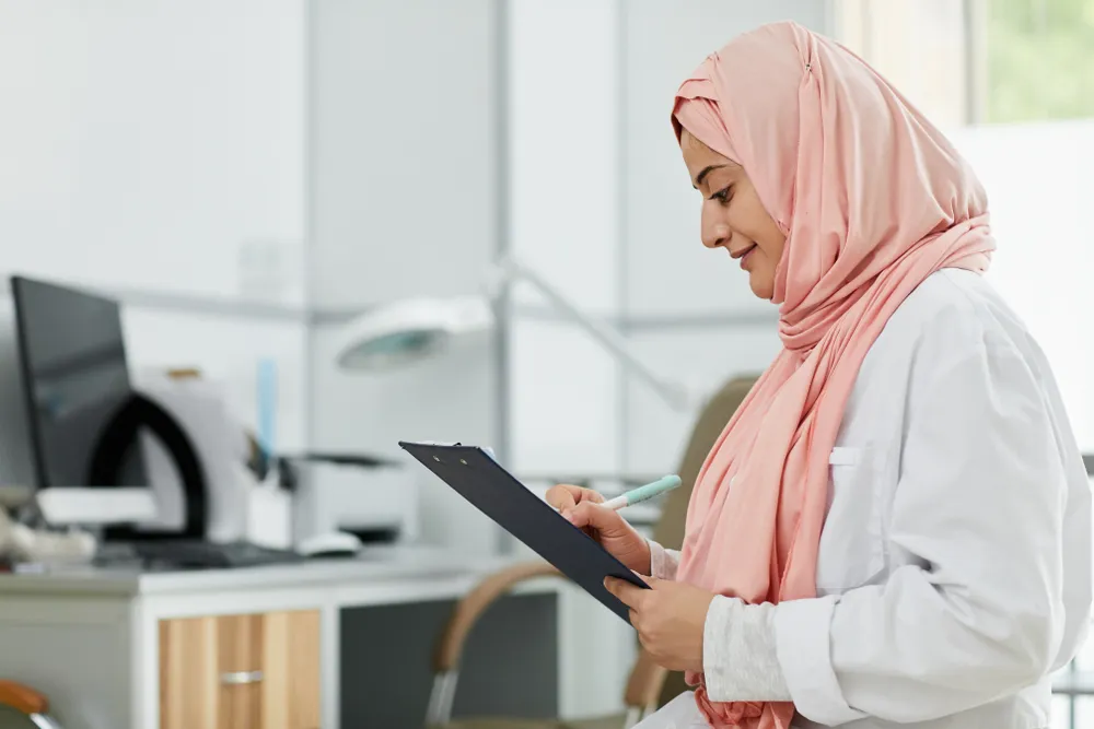female healthcare professional wearing a pink hijab, writing on a clipboard in a clinic.